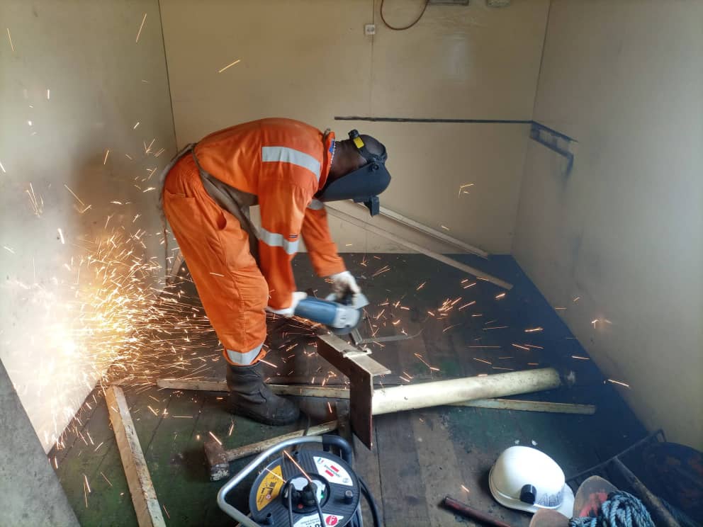Technicien PSG en tenue orange meulant une pièce métallique à l'intérieur d'un local industriel
